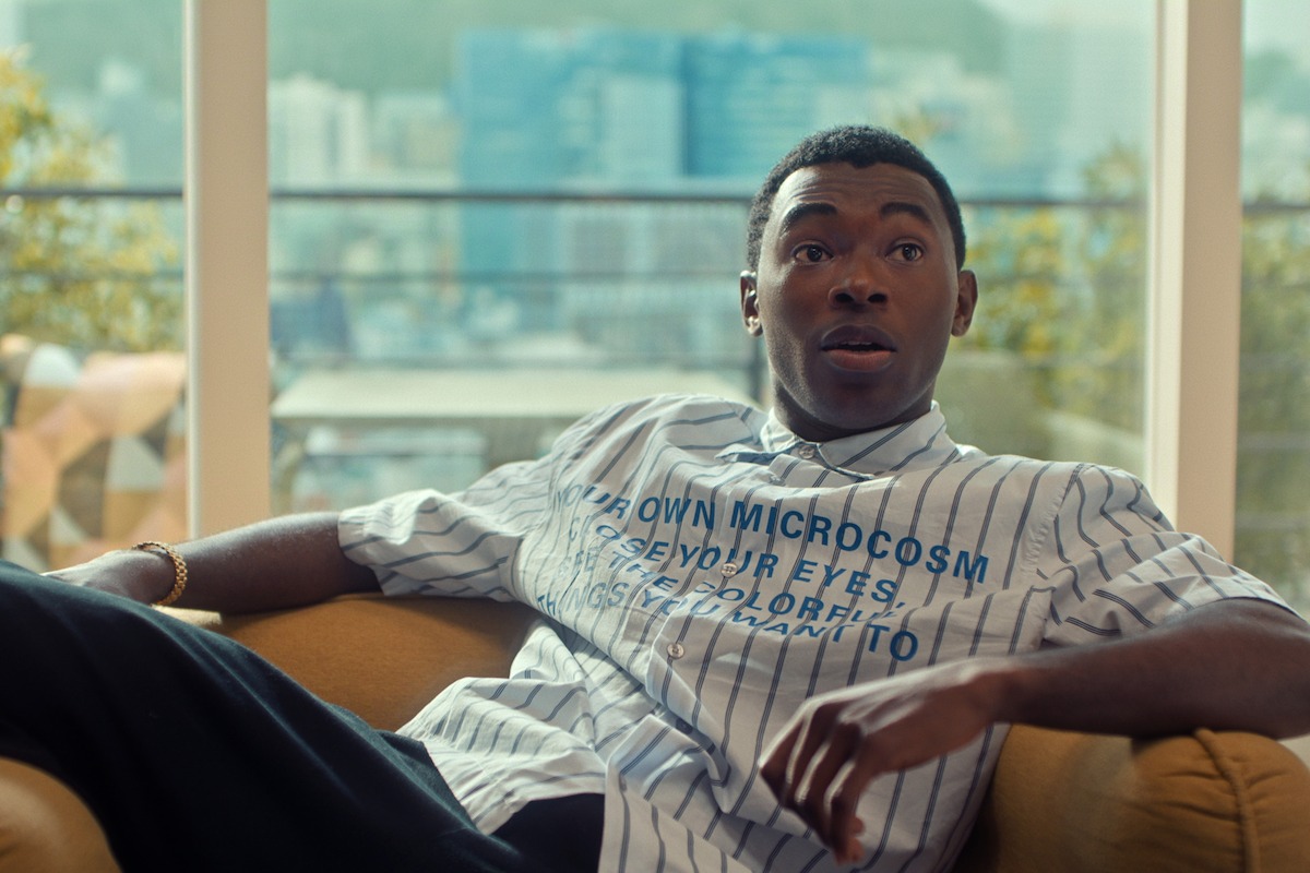 Man sitting on a yellow couch indoors, wearing a striped shirt with blue text, looking surprised. Large windows behind him show an urban cityscape with buildings and greenery in the background.