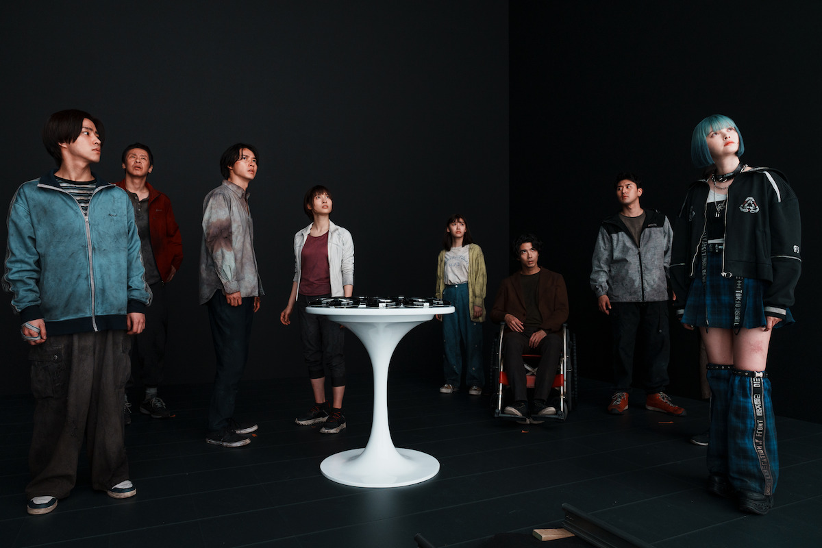 A group of people stand in an all-black space ion front of a small white table in an image from ‘Alice in Borderland’ Season 3.
