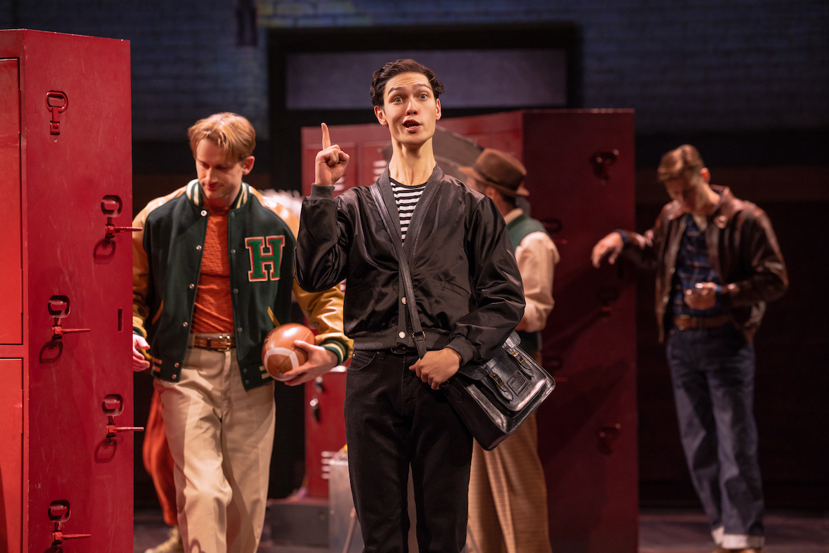 A young man in a black jacket gestures while standing in front of red lockers on a stage, surrounded by others in retro outfits, suggesting a school or locker room scene in a theatrical performance.