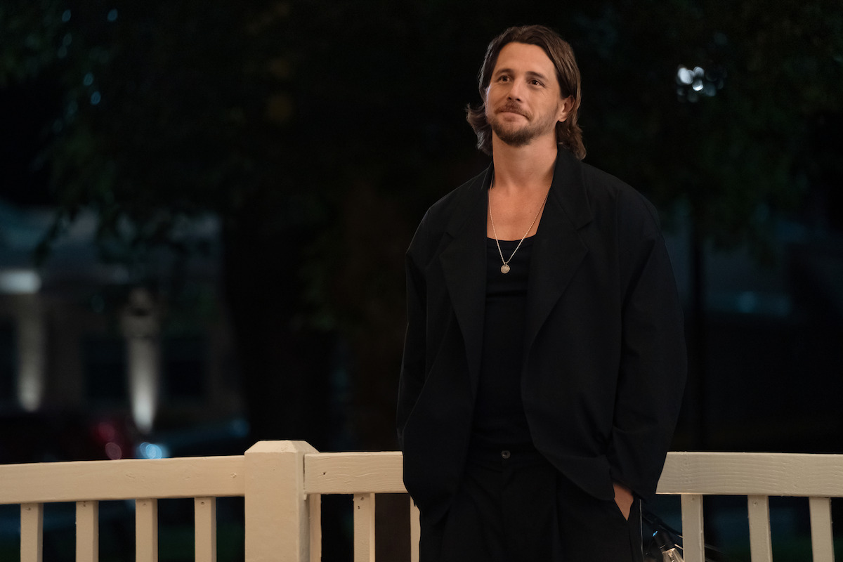 A man in a dark suit stands on a porch at night, leaning casually against a white railing with one hand in his pocket. Trees and dim outdoor lights are visible in the background.