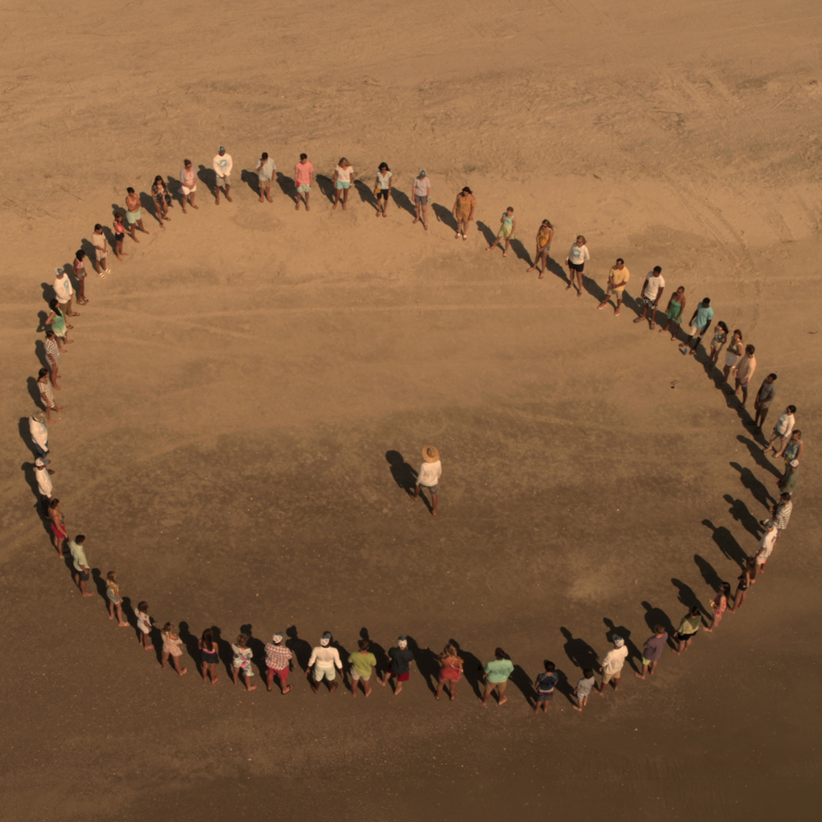 Um grupo de pessoas em pé em terreno arenoso forma um grande círculo em volta de uma pessoa que está no centro. A cena é vista de cima, em um ambiente amplo ao ar livre.