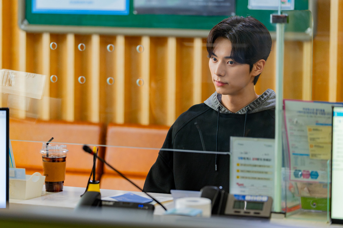 Young man sitting at a public service counter behind a glass barrier, with office supplies, documents, and an iced coffee on the desk, in a formal but calm indoor setting.