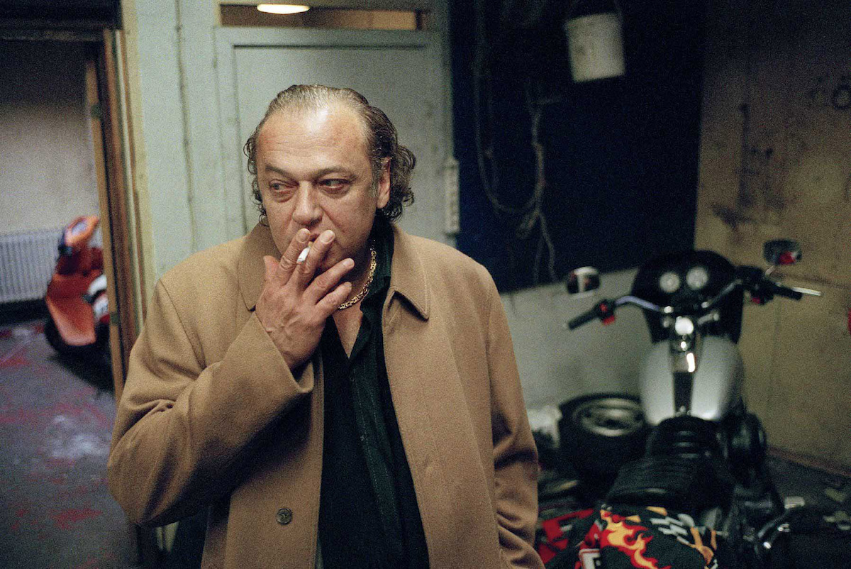 Milo (Zlatko Burić) wears a long camel coat and black shirt, and smokes a cigarette.