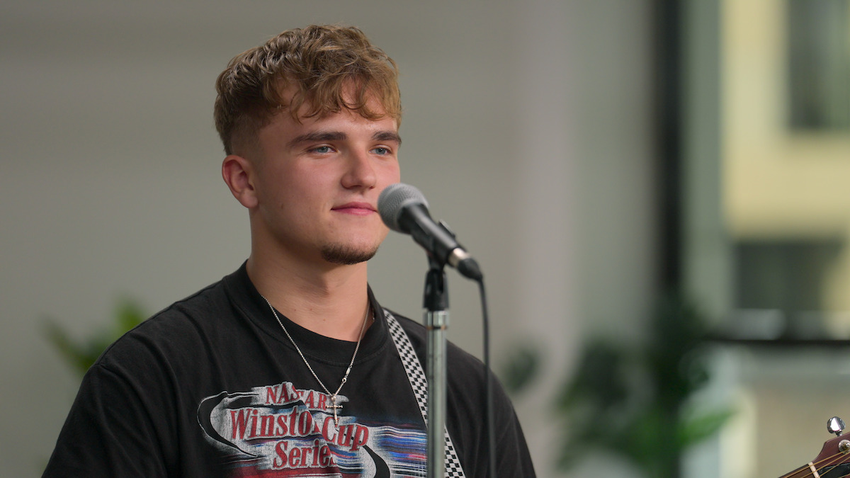 Young man stands indoors singing or speaking into a microphone, casual attire, guitar strap over shoulder, soft natural lighting, relaxed and focused mood, blurred background with plants and windows.