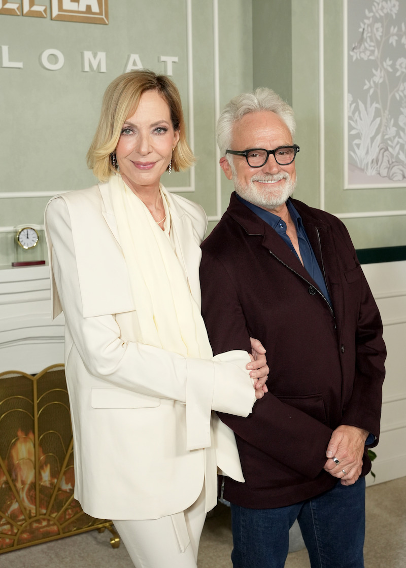 Two people pose together indoors in a stylish room with green walls, white decorative trim, and a fireplace. The person on the left wears a white suit, and the person on the right wears glasses and a dark jacket.