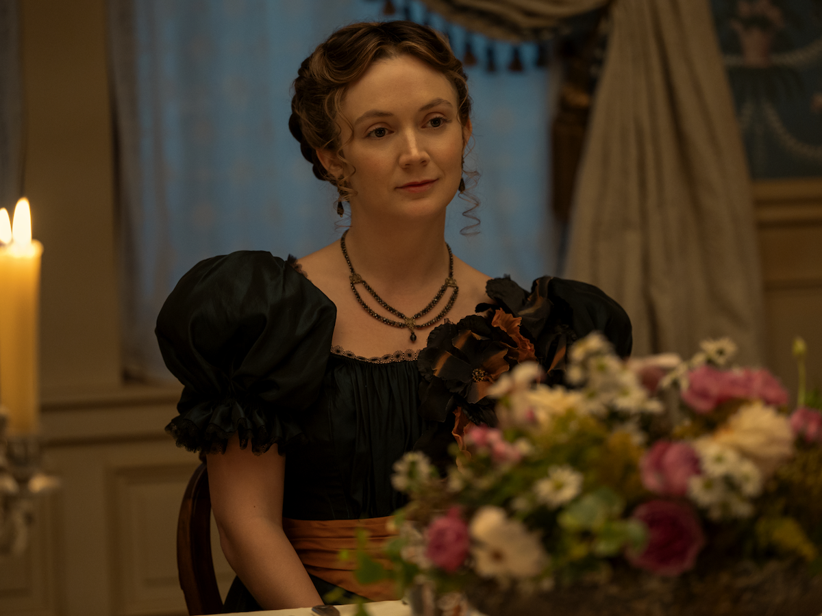 A woman in period costume sits at a candlelit dinner table with a floral centerpiece in an elegantly decorated room, featuring classic drapes and soft lighting.