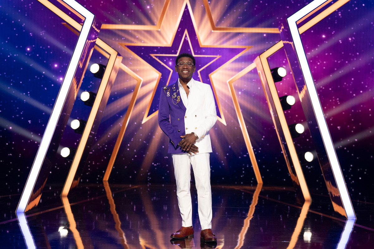 Spud Howarrd stands center stage, with a large illuminated star backdrop, stage lights, and a sparkling purple background, suggesting a talent show or competition environment.