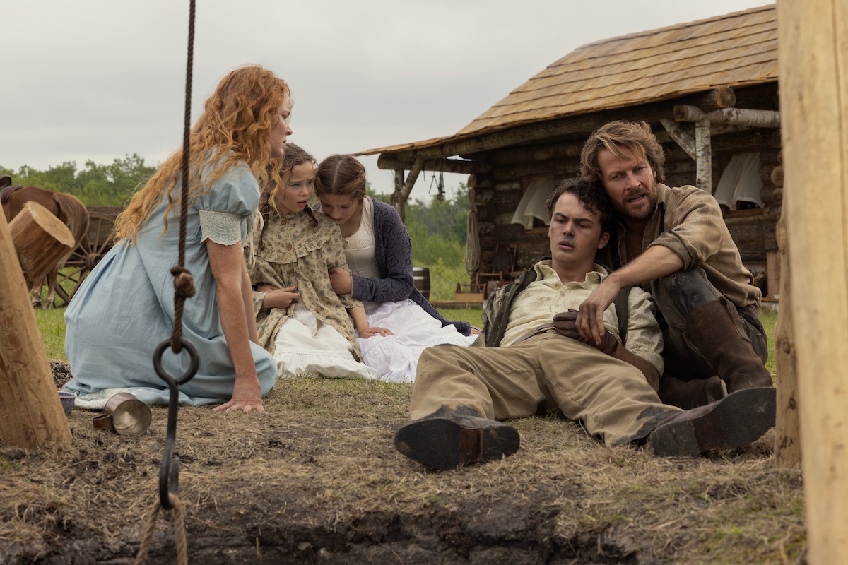 Five people in period clothing sit on the ground next to a well in a rural, rustic setting with a wooden cabin in the background, appearing distressed or comforting one another.