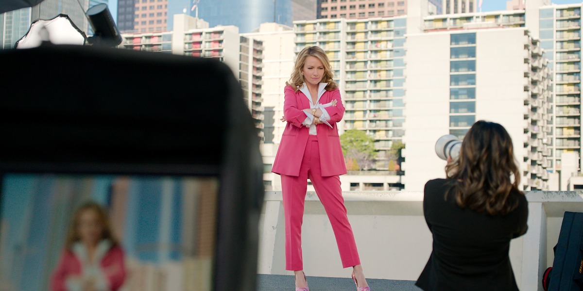 Woman in a pink suit poses for a photographer on a rooftop with tall city buildings in the background, camera and equipment visible in the foreground.