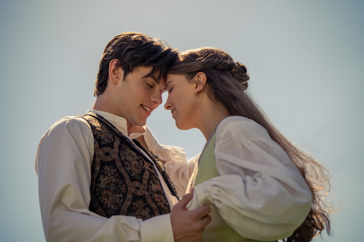 A young couple in vintage clothing stands close together outdoors, gently touching foreheads and smiling, with a soft blue sky background and warm natural light creating a romantic, serene atmosphere.