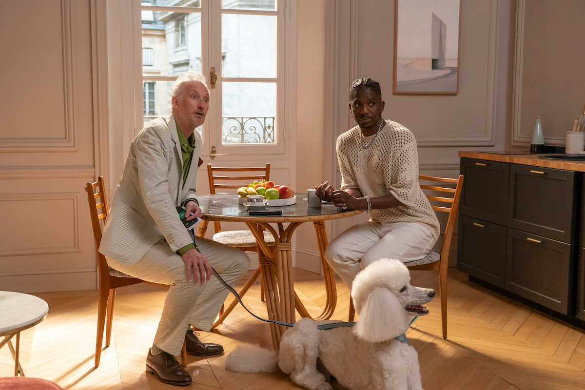 Two men sitting at a round wooden table in a bright, elegant apartment with a window, fruit bowl, and modern decor. A white poodle on a leash sits on the floor beside them.