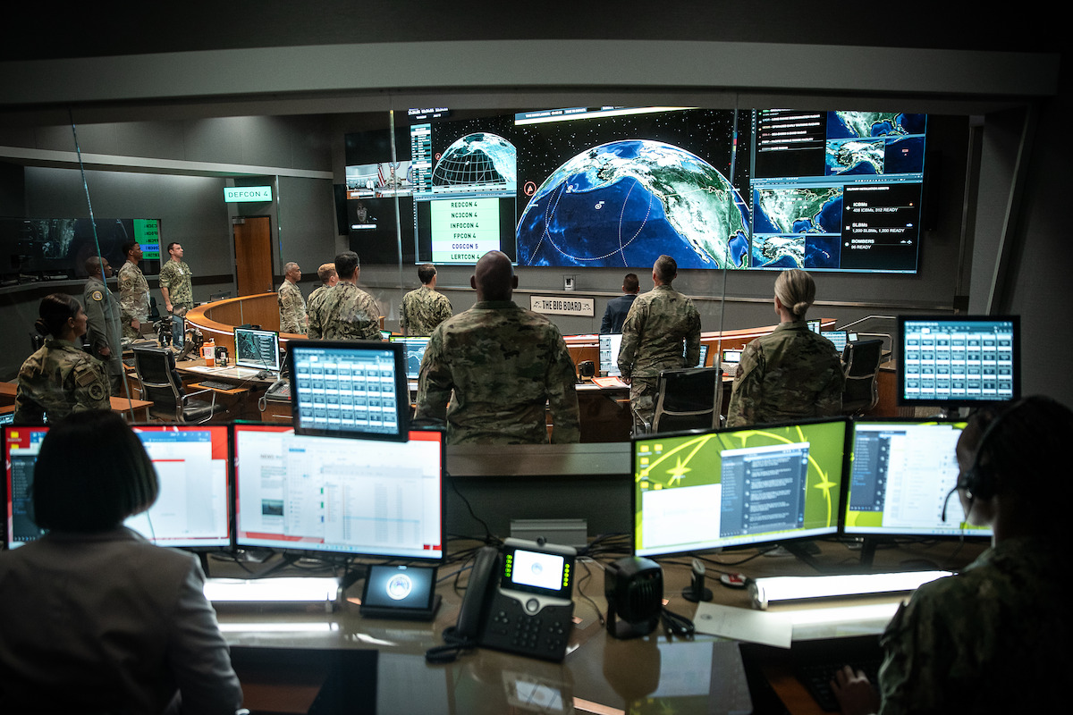 Military personnel in a high-tech control room monitor multiple screens with satellite and earth data. The atmosphere is serious and focused, with rows of computers and large digital displays showing global strategic information.