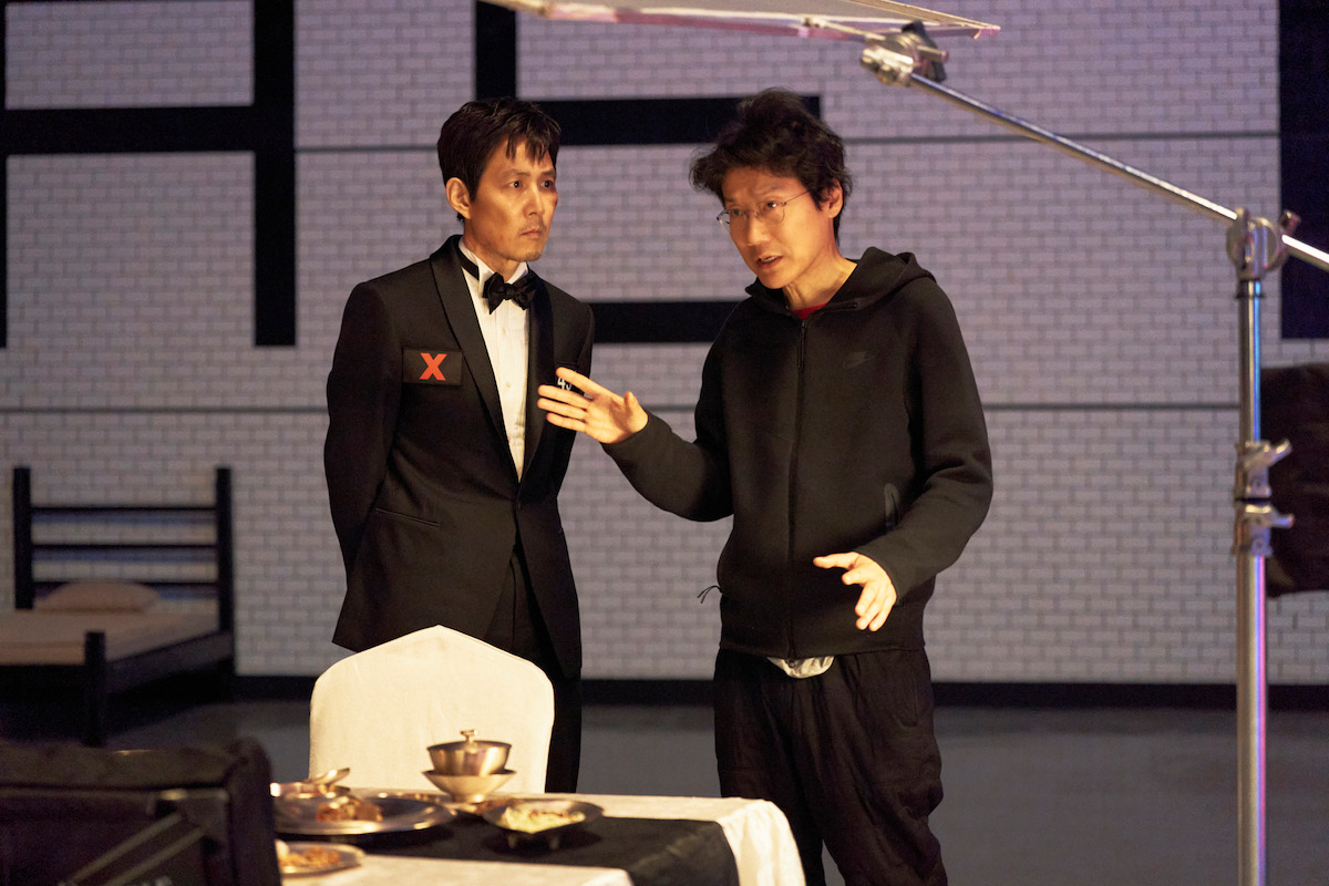 Two men are on a set with a dining table and filming equipment. One wears a tuxedo, standing by a chair with dishes on a table, while the other appears to explain something, gesturing with his hands in a modern, indoor environment.