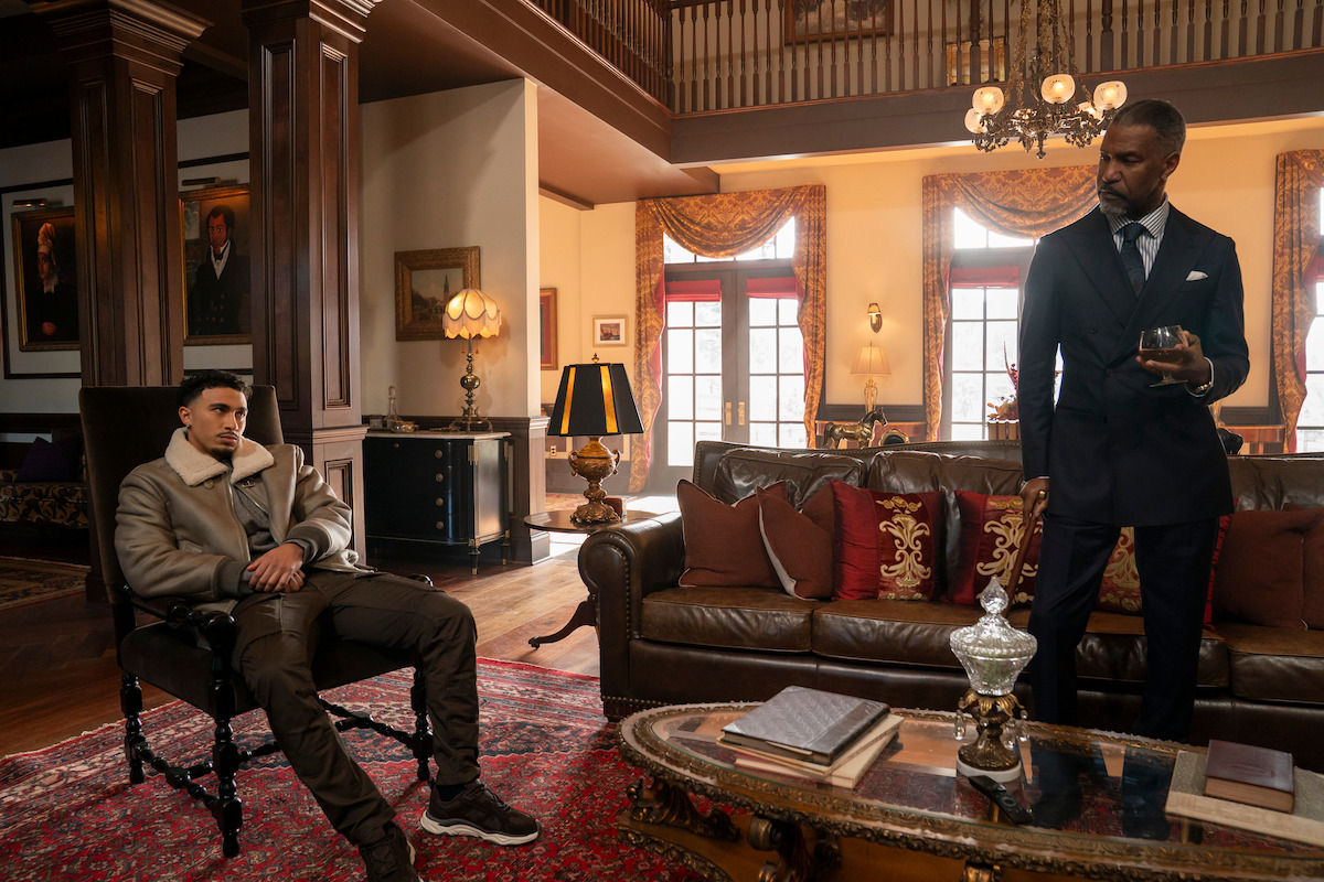 Two men in a luxurious living room; one is seated casually in a chair while the other stands by a sofa holding a drink. The room has elegant décor, large windows, wooden columns, ornate furniture, warm lighting, and a red rug.