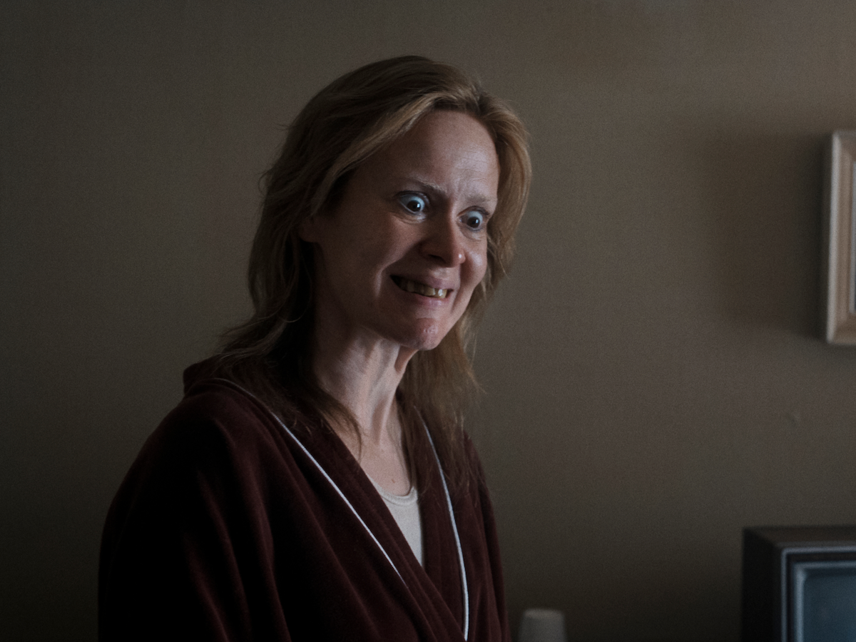 Woman with a strained smile standing indoors in dim lighting, wearing a maroon robe. The background includes a brown wall and part of a picture frame, creating a tense and unsettling atmosphere.