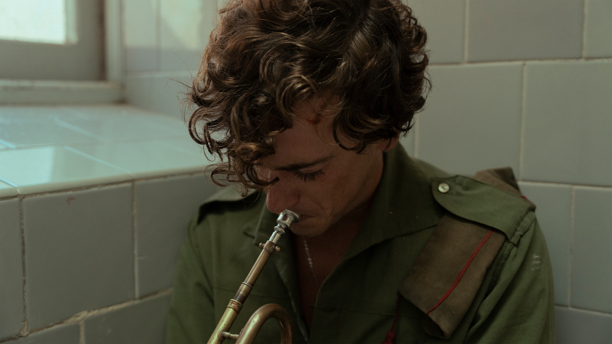 A young man in a green military uniform sits in a tiled room, head bowed as he holds a trumpet to his lips, appearing thoughtful or somber in a softly lit, quiet environment.