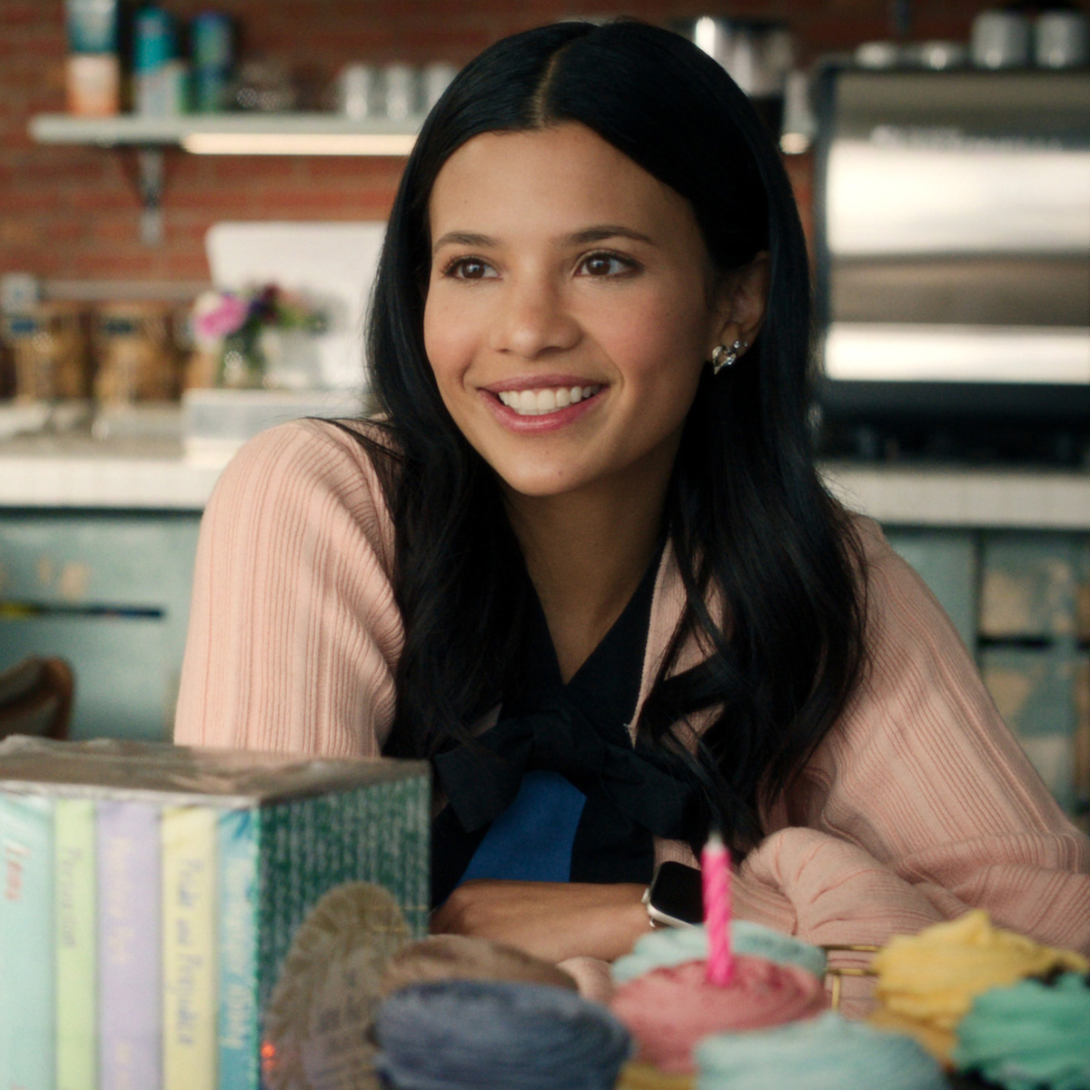 A smiling woman with dark hair sits at a table with cupcakes, yarn, and books in a cozy kitchen or café with a coffee machine and shelves in the background, creating a warm and inviting atmosphere.