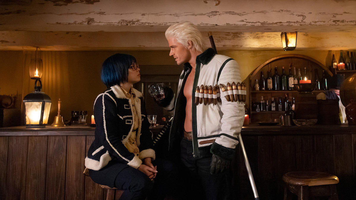 Two people in elaborate outfits have a serious conversation at a rustic bar; one sits on a stool while the other stands, holding a drink. The background shows shelves with bottles, warm lighting, and wood furnishings.