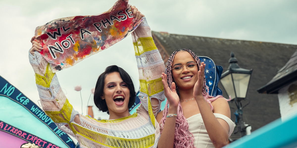 Two smiling people outdoors at a pride parade, one holding a colorful sign that says "We are not a phase," with festive decorations and a streetlamp in the background.