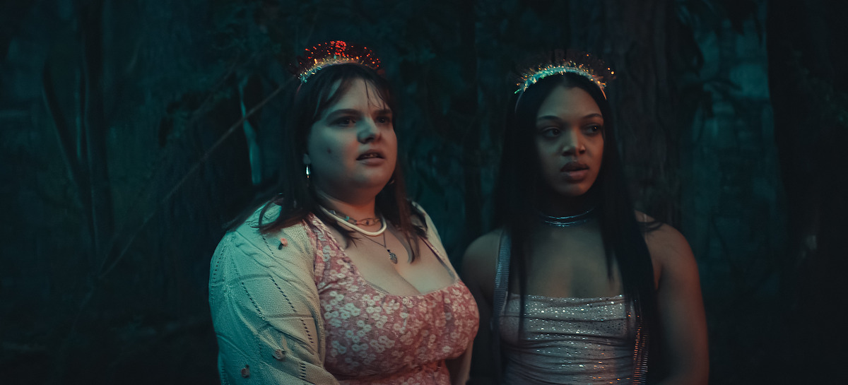Two teenage girls in party dresses and tiaras stand close together in a dark, wooded area, looking ahead with anxious, uncertain expressions.