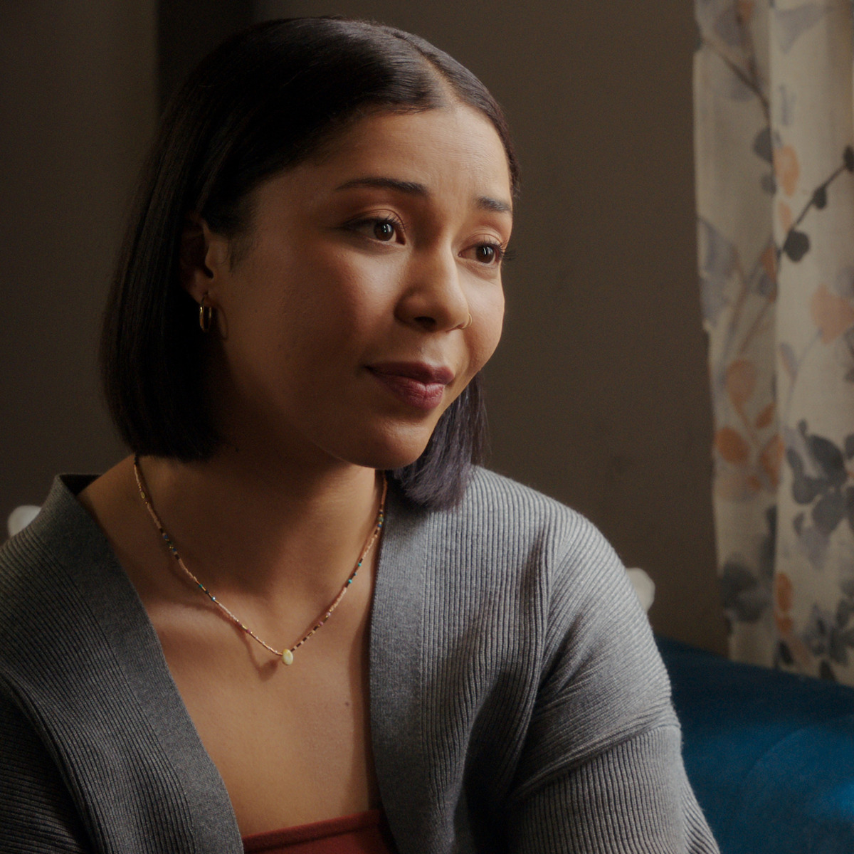 Woman with straight dark hair and gold jewelry sits indoors on a blue chair, wearing a gray sweater and rust-colored top. Light from a window with floral curtains gently illuminates her, creating a warm and calm atmosphere.