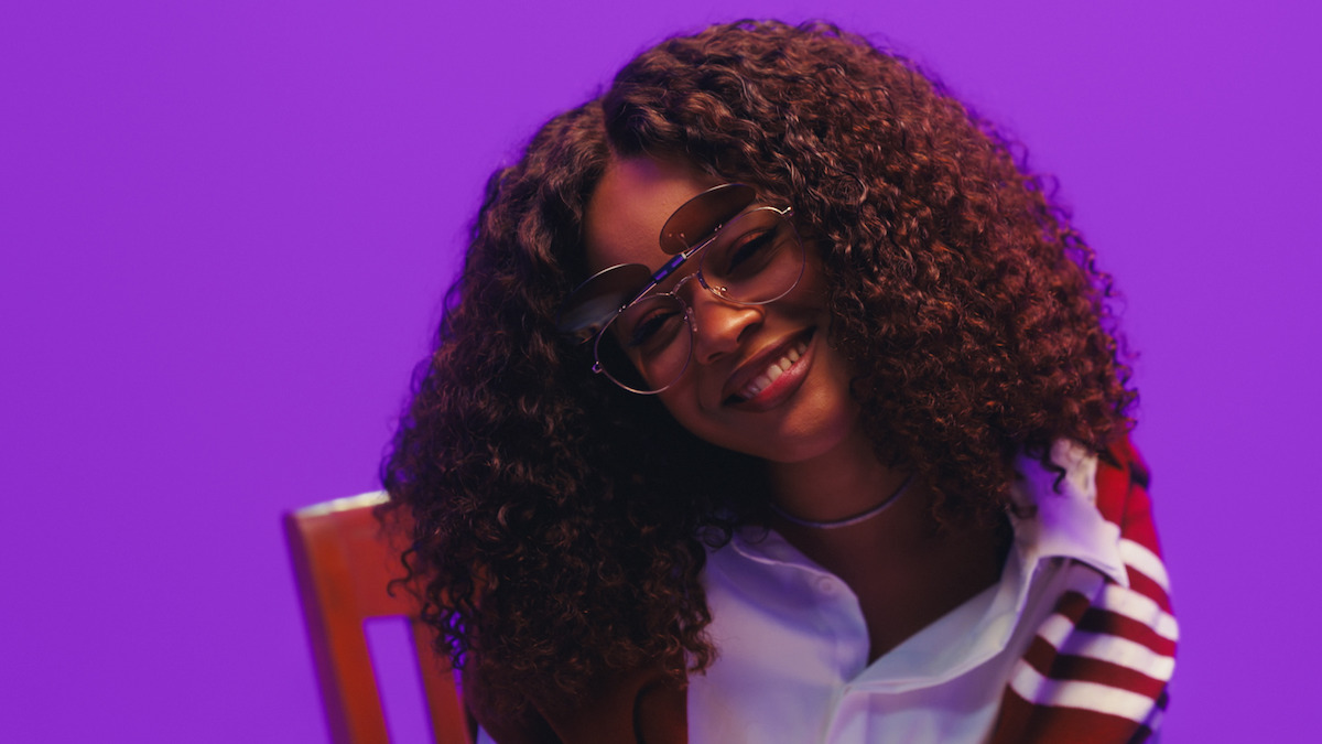 Smiling young woman with curly hair and sunglasses sitting on a chair in front of a purple background, wearing a white shirt and striped jacket.