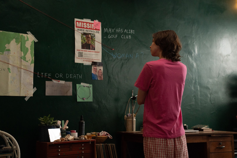 Teenager in pink shirt stands in bedroom, studying a green wall covered with clues, photos, missing person poster, map, and handwritten notes for a mystery investigation, suggesting a detective or suspense scenario.