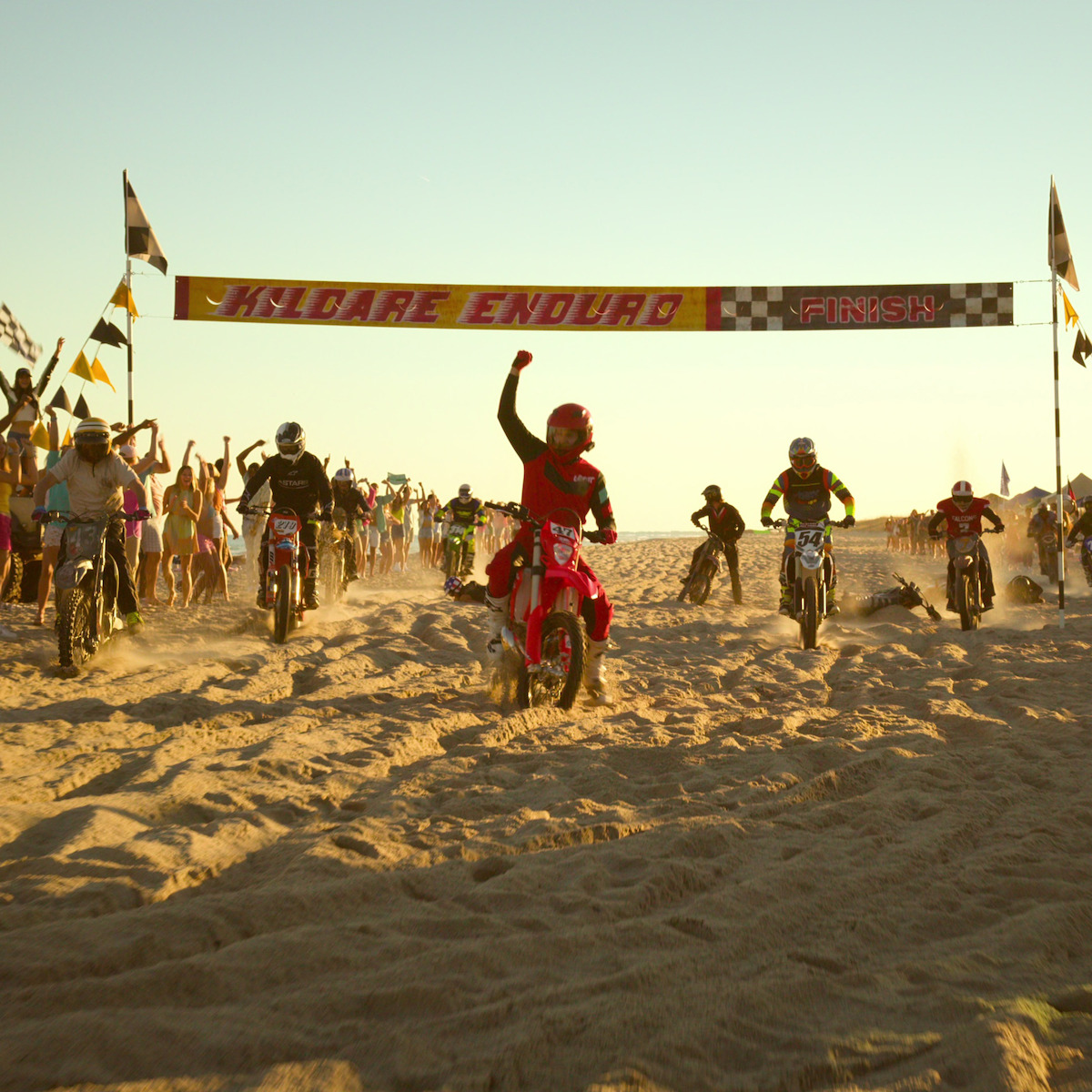Motociclistas disputam uma corrida na areia da praia, com uma multidão torcendo e bandeiras decorando o ambiente durante o pôr do sol. Um participante ergue o punho, triunfante, ao cruzar a linha de chegada.