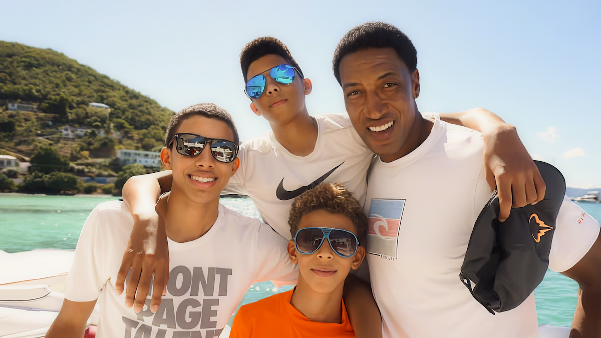 Four people smiling and posing together on a boat by turquoise water, with hills and houses in the background on a sunny day.