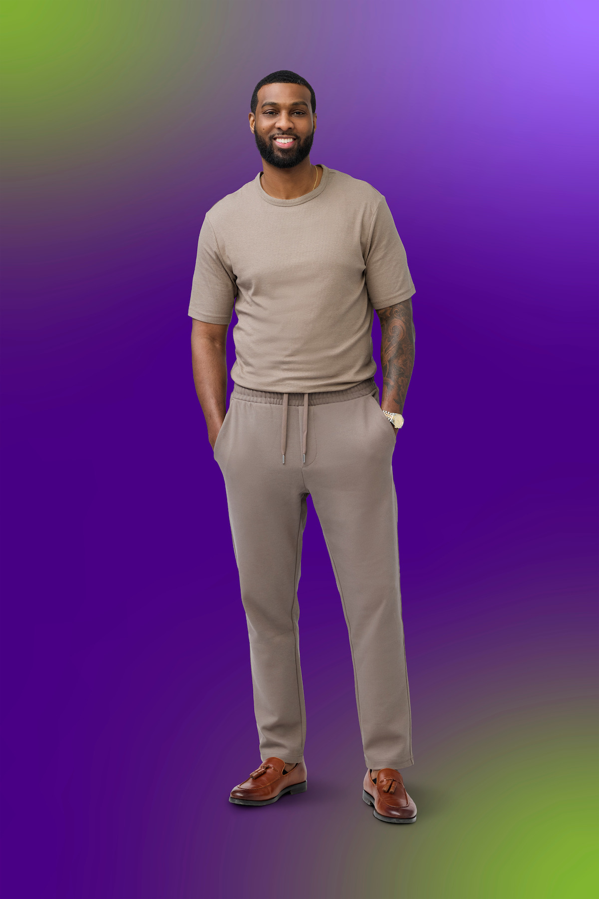 A man stands smiling in casual beige clothes with brown shoes, hand in pocket, against a vibrant purple and green gradient background, creating a modern, relaxed atmosphere.
