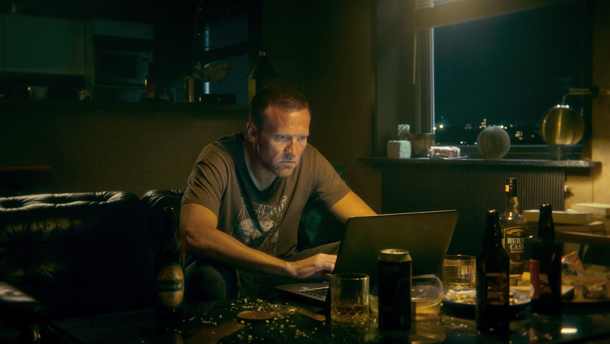 A man sits in a dimly lit, cluttered room at night, focusing intently on his laptop. The environment feels tense and isolated, with bottles and plates scattered around a table, suggesting loneliness or stress.