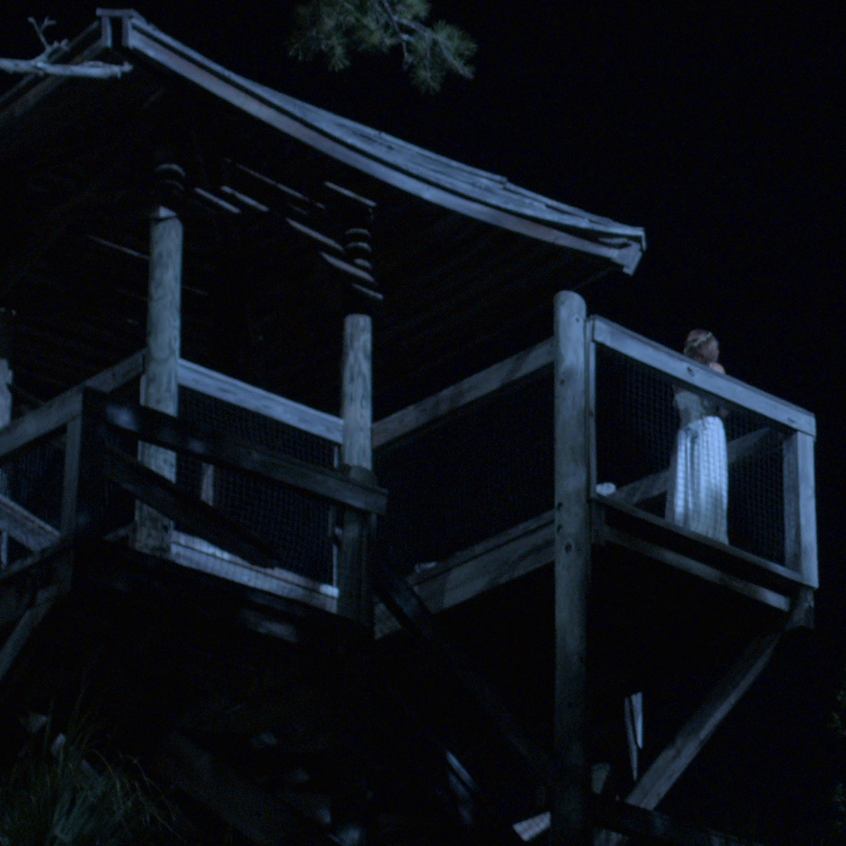 Uma pessoa de vestido branco está em pé no balcão superior de uma torre de vigia à noite, cercada pela escuridão e iluminada parcialmente pelo luar.