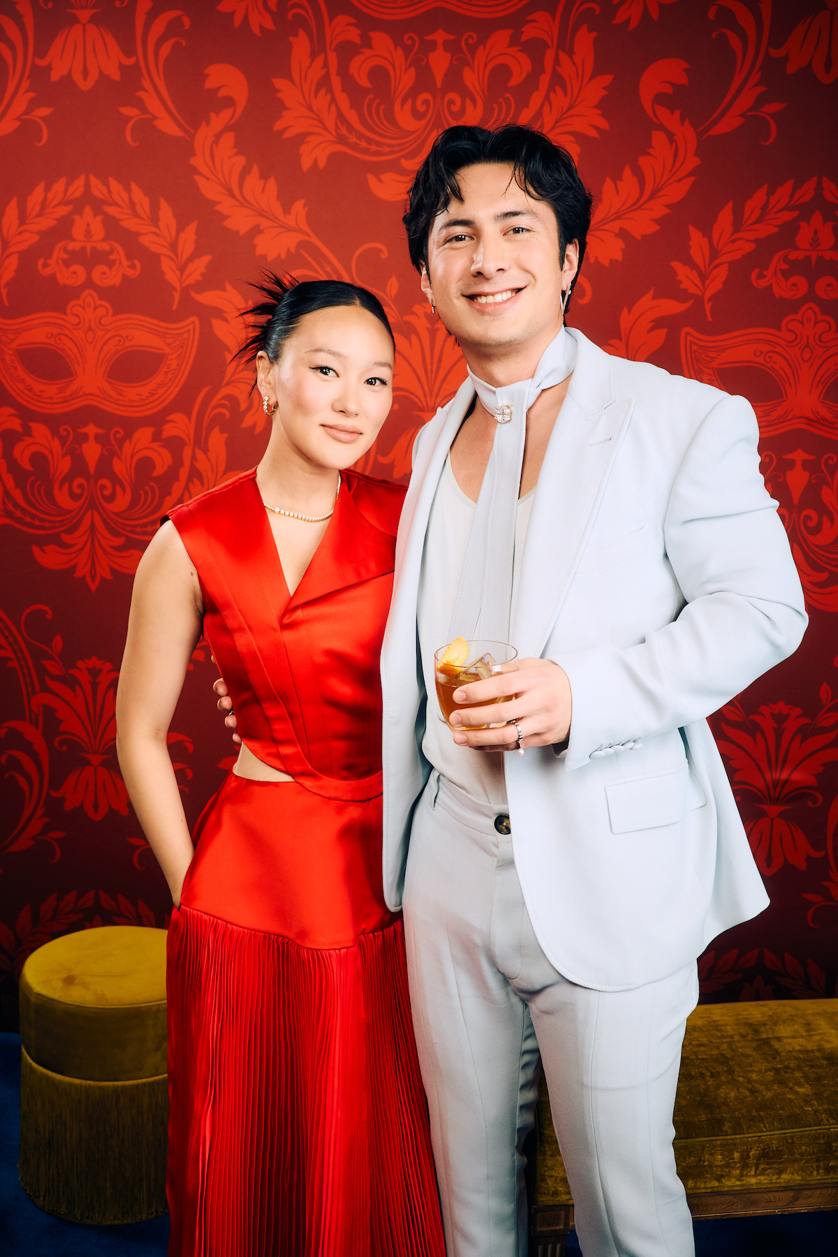 Duas pessoas posam juntas em um evento formal. Uma delas usa um vestido vermelho, enquanto a outra veste um traje claro e segura uma bebida. Elas estão diante de uma parede vermelha, estampada com detalhes dourados e móveis decorativos.