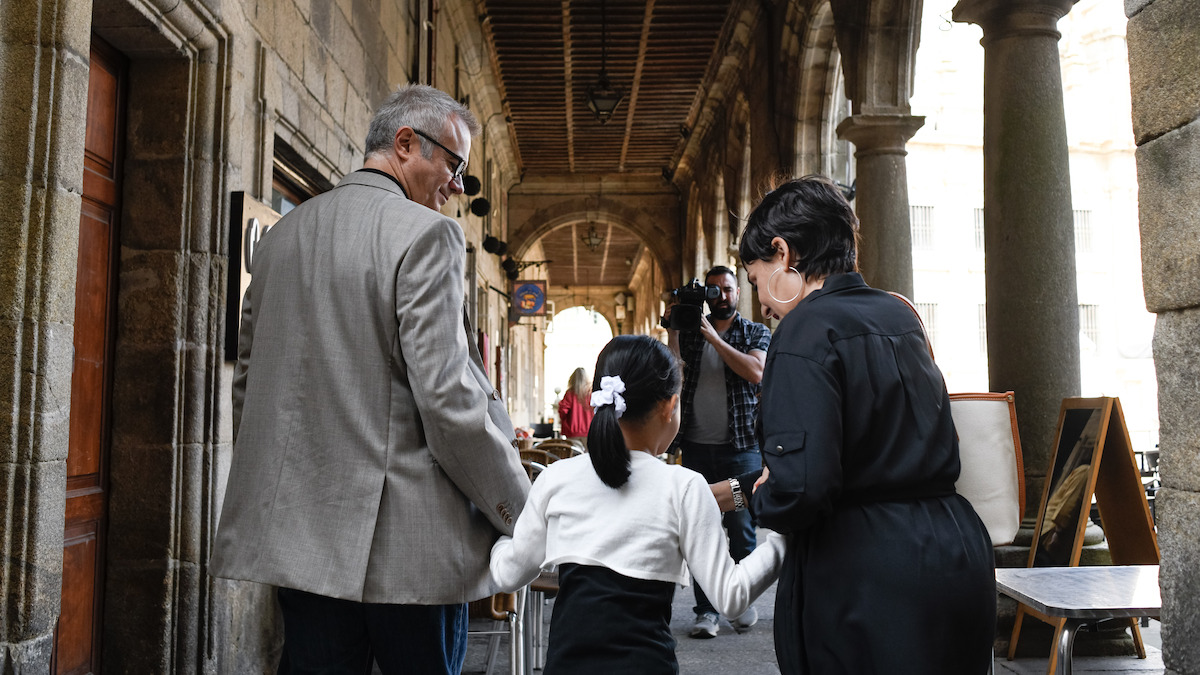 In a still image from the series ‘The Asunta Case,’ Tristán Ulloa as Alfonso and Candela Peña as Rosario hold Iris Wu as Asunta’s hand while the trio walk down the sidewalk in Santiago, Spain.
