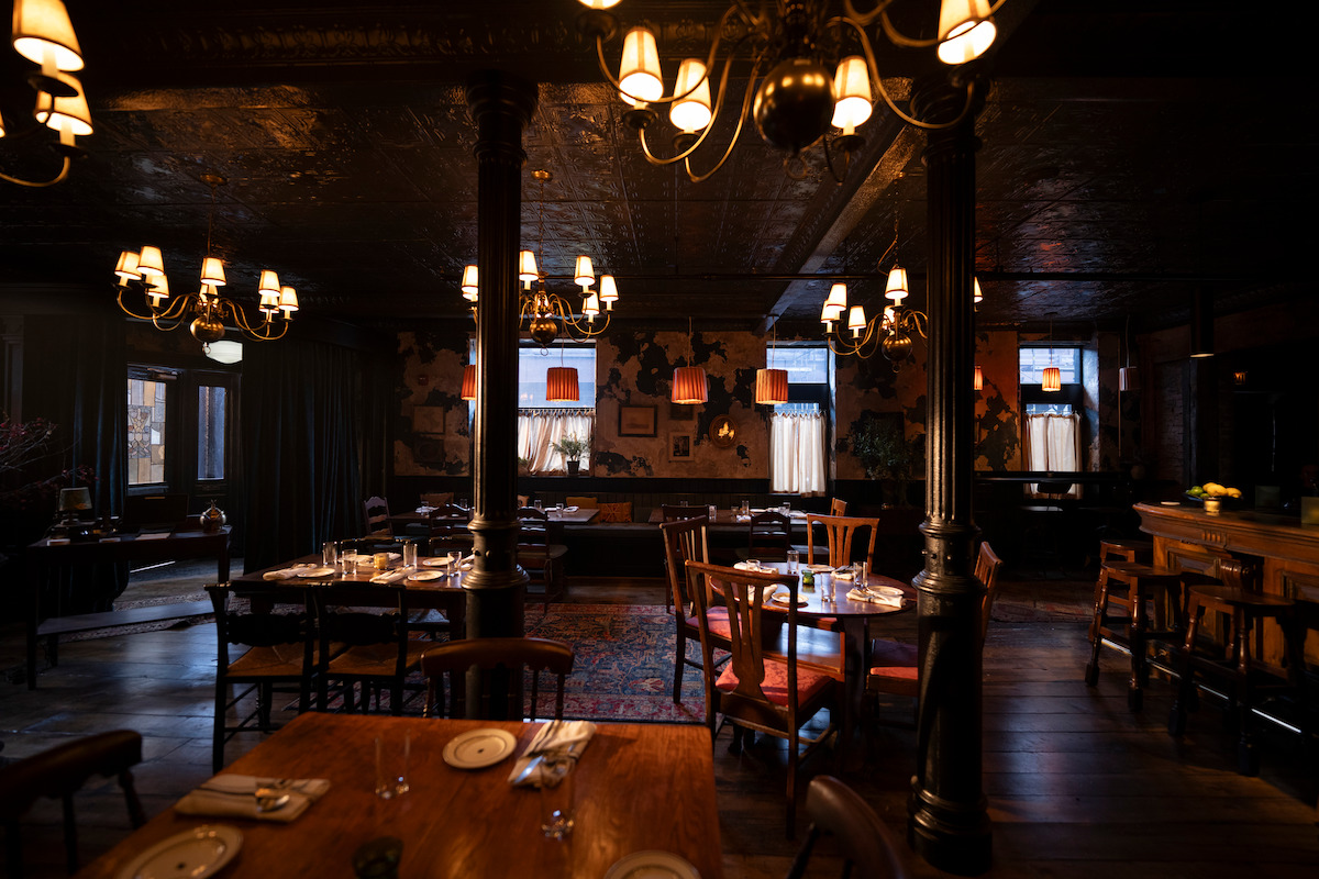 Cozy, dimly lit restaurant interior with wooden tables, vintage chandeliers, large pillars, set dining tables, and rustic decor, creating a warm and inviting atmosphere.