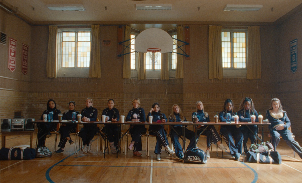 A group of girls in athletic uniforms sit behind a long table in a gymnasium, with sports bags on the floor, under a basketball hoop and surrounded by tall windows and wall banners.