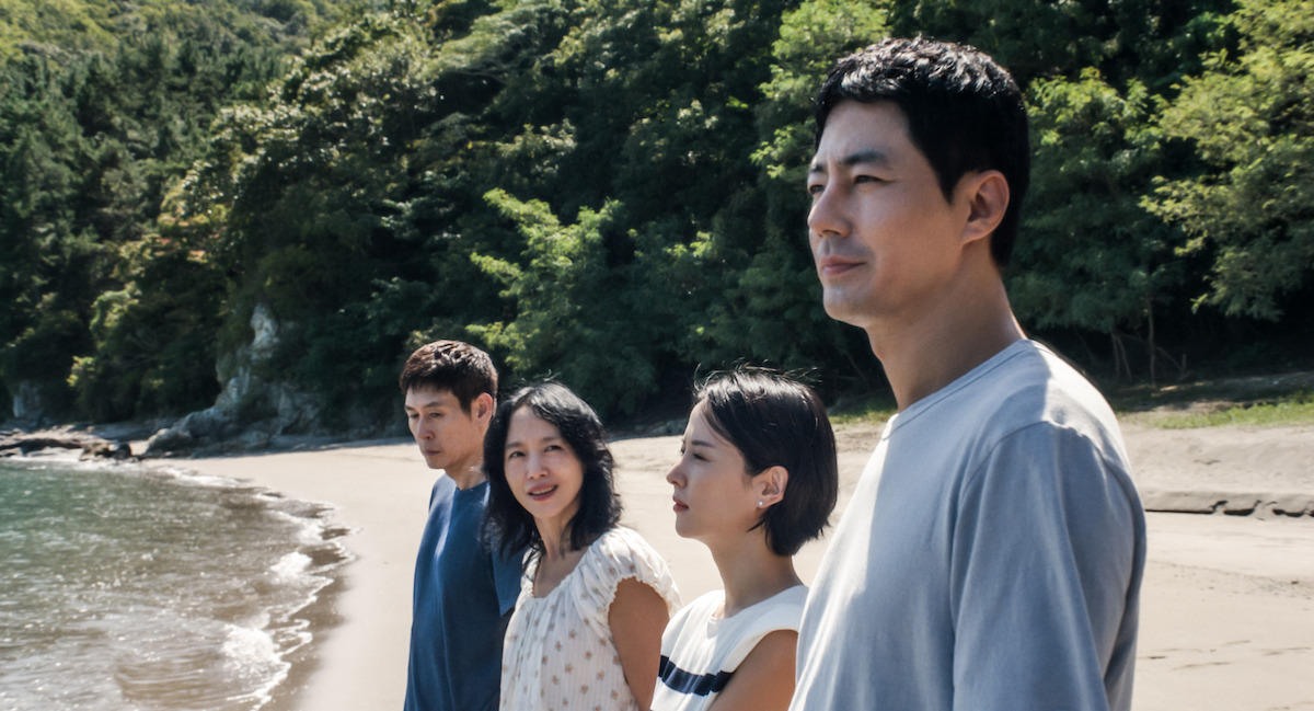 Four people stand on a sandy beach near the water’s edge, facing slightly toward the camera, with green forested hills and trees in the background under daylight.
