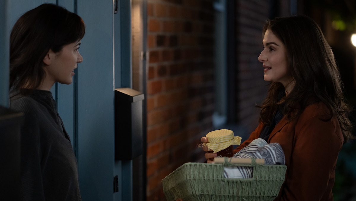 Two women talking at a doorway at night, one holding a green basket with folded towels and a container, standing outside near a brick wall and blue door.