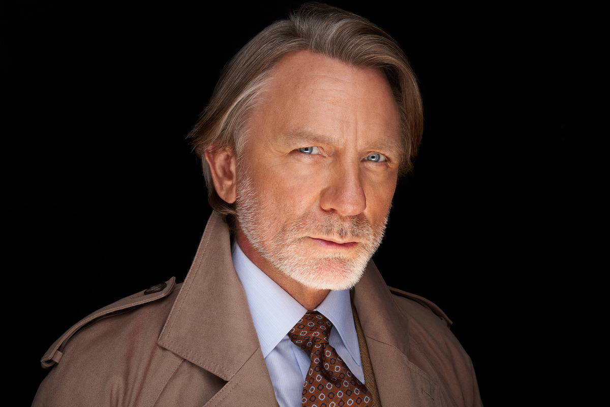 Serious older man with light hair and beard in a trench coat, dress shirt, and patterned tie, posed against a dark background with dramatic lighting, creating an intense and focused mood.