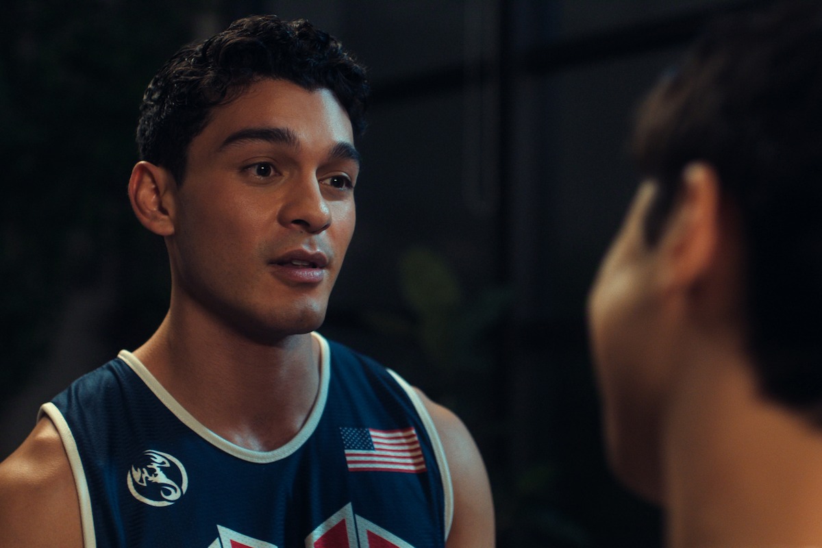 Young man in a basketball jersey with USA and dragon patches talks to another person indoors at night with a blurred background and some plants visible.