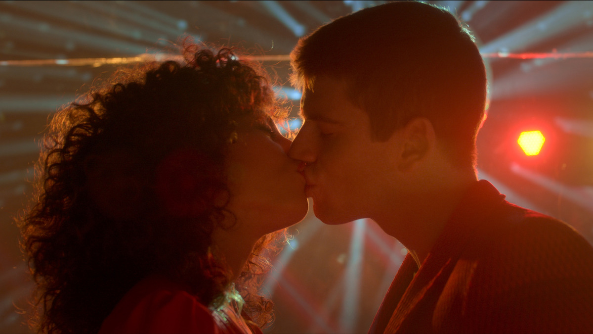 Two people kissing in a dramatic, colorful setting with bright stage lights and beams radiating behind them, suggesting a romantic moment at a party or nightclub.