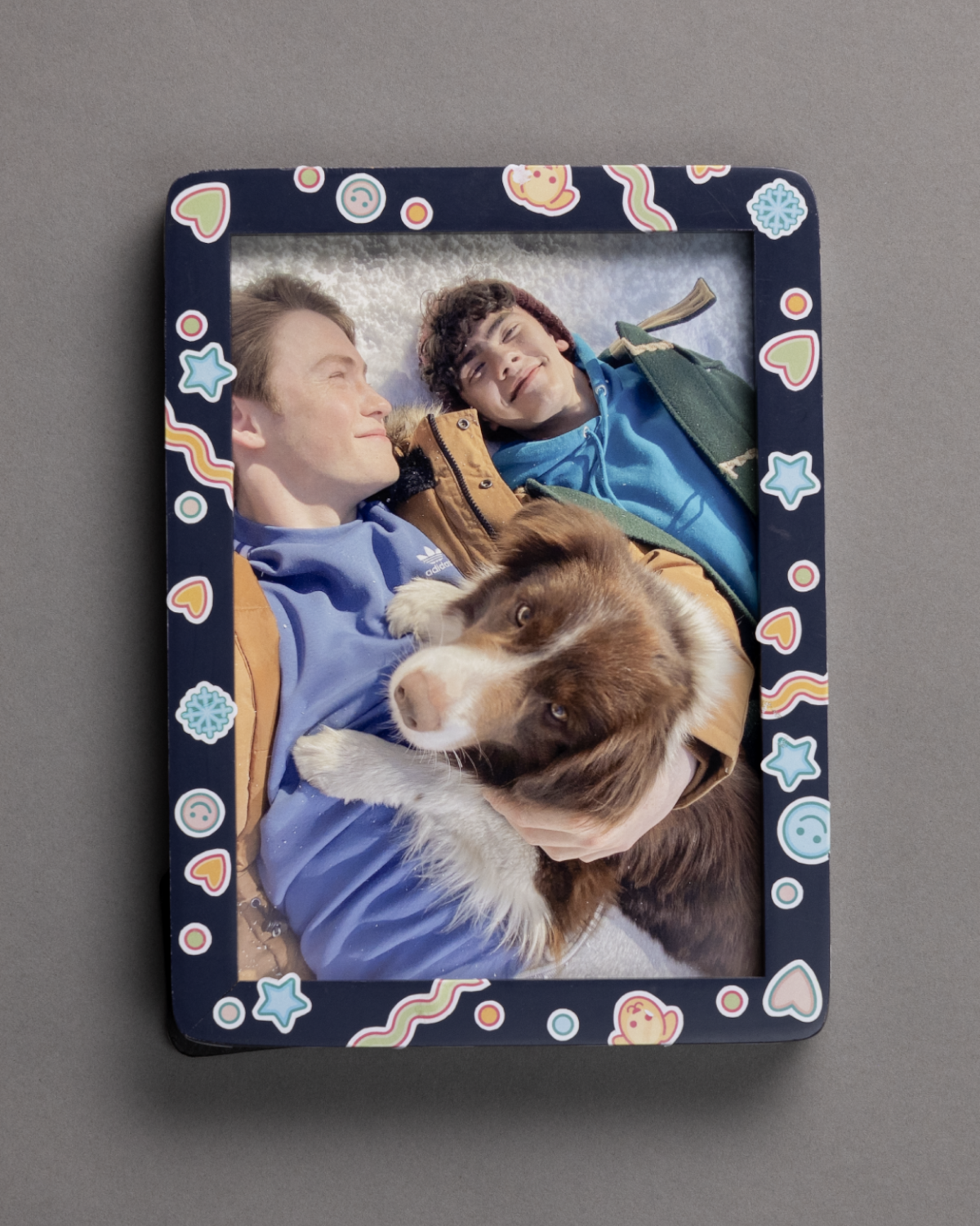 Two people lie on the ground outdoors, smiling at each other, with one holding a brown and white dog. Photo is in a dark, playful frame decorated with hearts, stars, and smiley faces, resting on a plain gray surface.