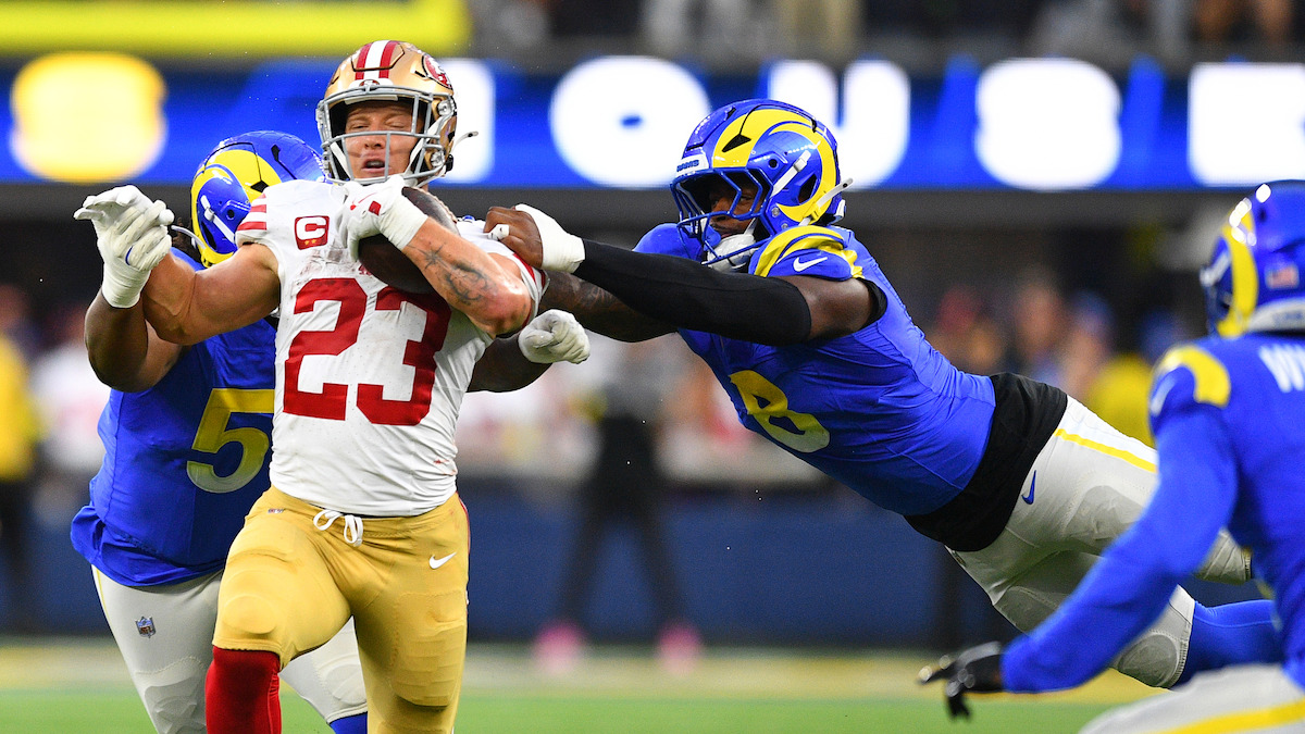 Football player in red and gold uniform running with the ball, being tackled by two players in blue and yellow uniforms on a professional field during a game. Stadium lights and blurred audience in the background.