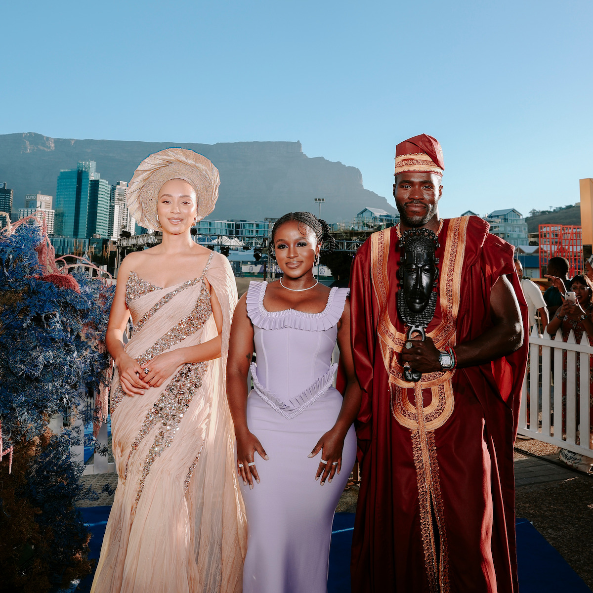 Three people in elegant attire pose outdoors at an event, with city buildings and a mountain in the background under a clear blue sky.