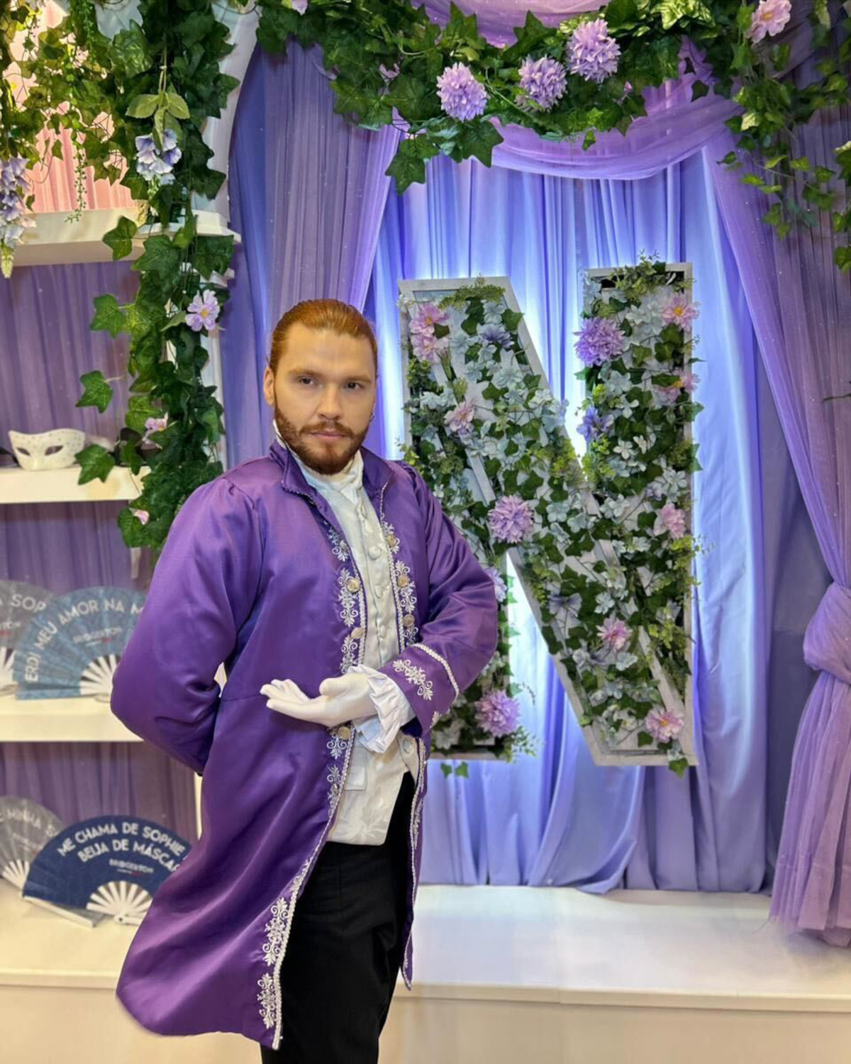 Pessoa com traje roxo no estilo do século 18 posa diante de uma grande letra N decorada com hera e flores, com tecidos roxos e decorações florais, em um cenário lúdico elegante.