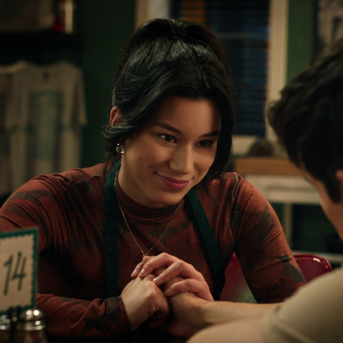 A young woman sitting in a cozy café smiles warmly and clasps hands with someone across the table, suggesting a caring, intimate moment. The background shows a casual dining setting with soft lighting and decor.