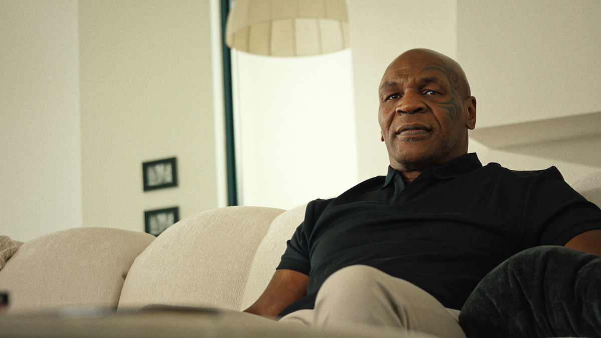 Man sitting on a light-colored couch in a modern, bright living room with minimal decor, soft lighting, and neutral tones, appearing relaxed and contemplative.