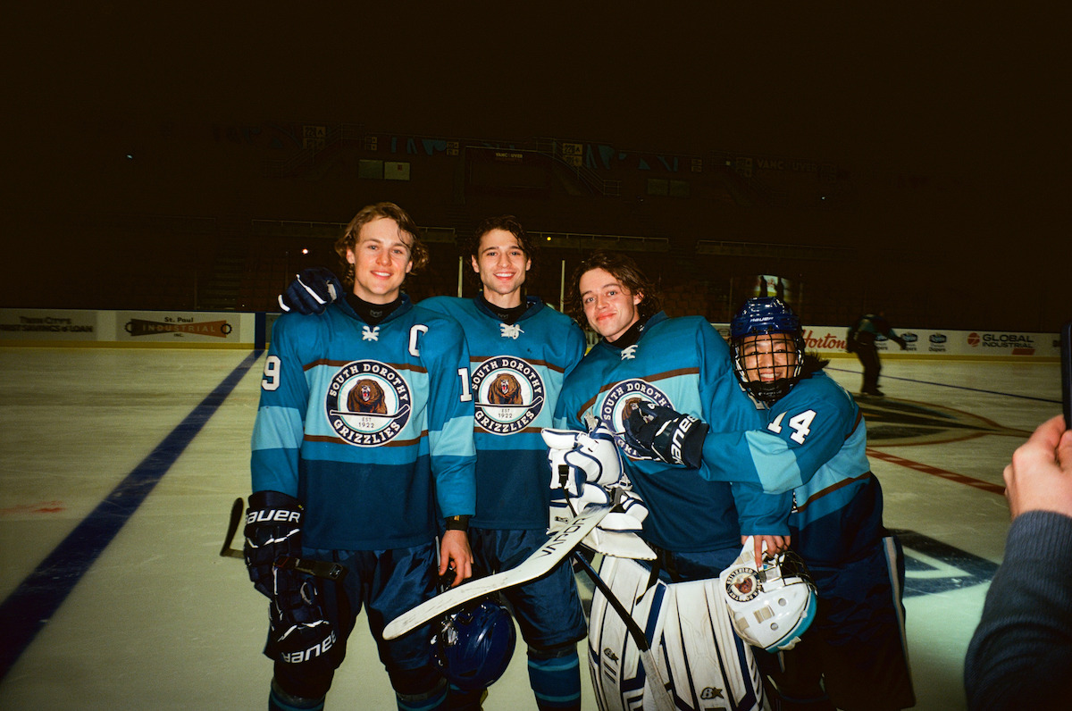Four hockey players in blue uniforms posing together on an ice rink, smiling for a photo. The rink is indoors with boards and advertisements visible in the background, suggesting a team game or practice session.