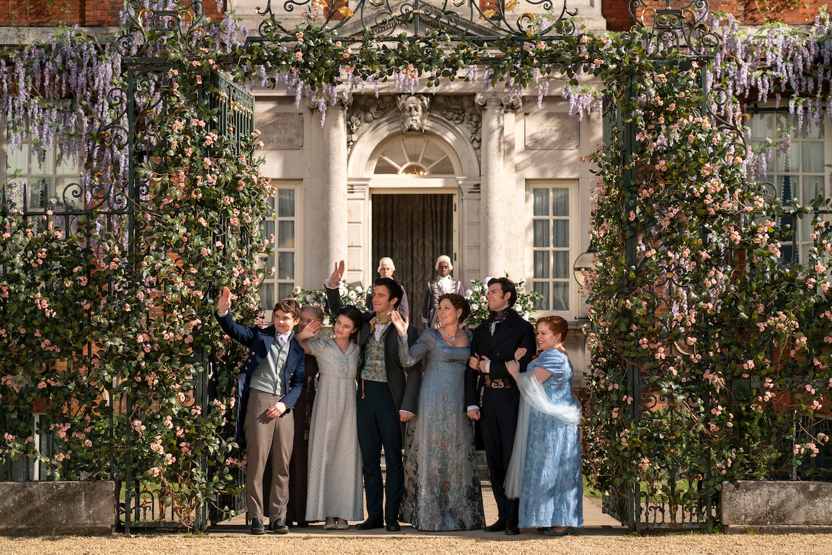 Six people in Regency-era clothing stand together outside a grand estate with flowers and vines, posing at an ornate gate with a historic mansion in the background on a sunny day.