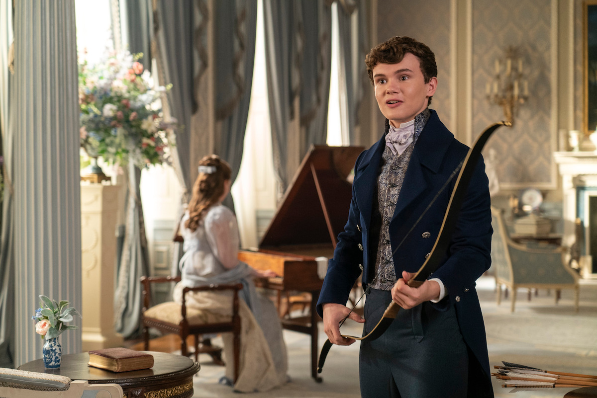 Young man in Regency-era clothing holding a bow in an elegant, sunlit room with classic decor; woman sits by a grand piano, floral arrangement and ornate furnishings in the background, suggesting a historical or period drama setting.